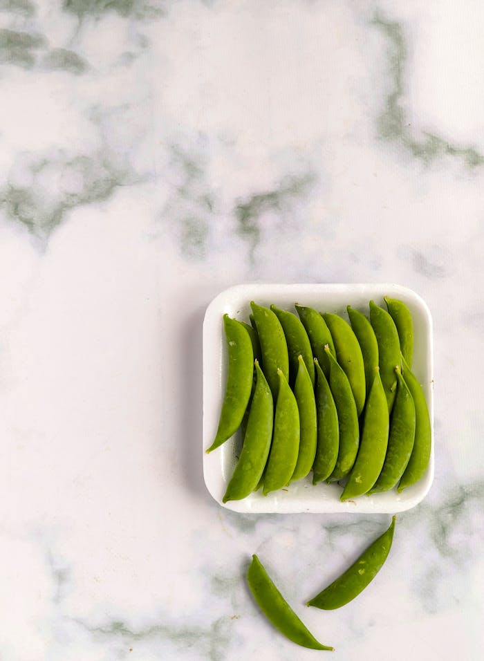 Fresh sugar snap peas neatly arranged on a white tray against a marble background. Perfect for healthy recipes.